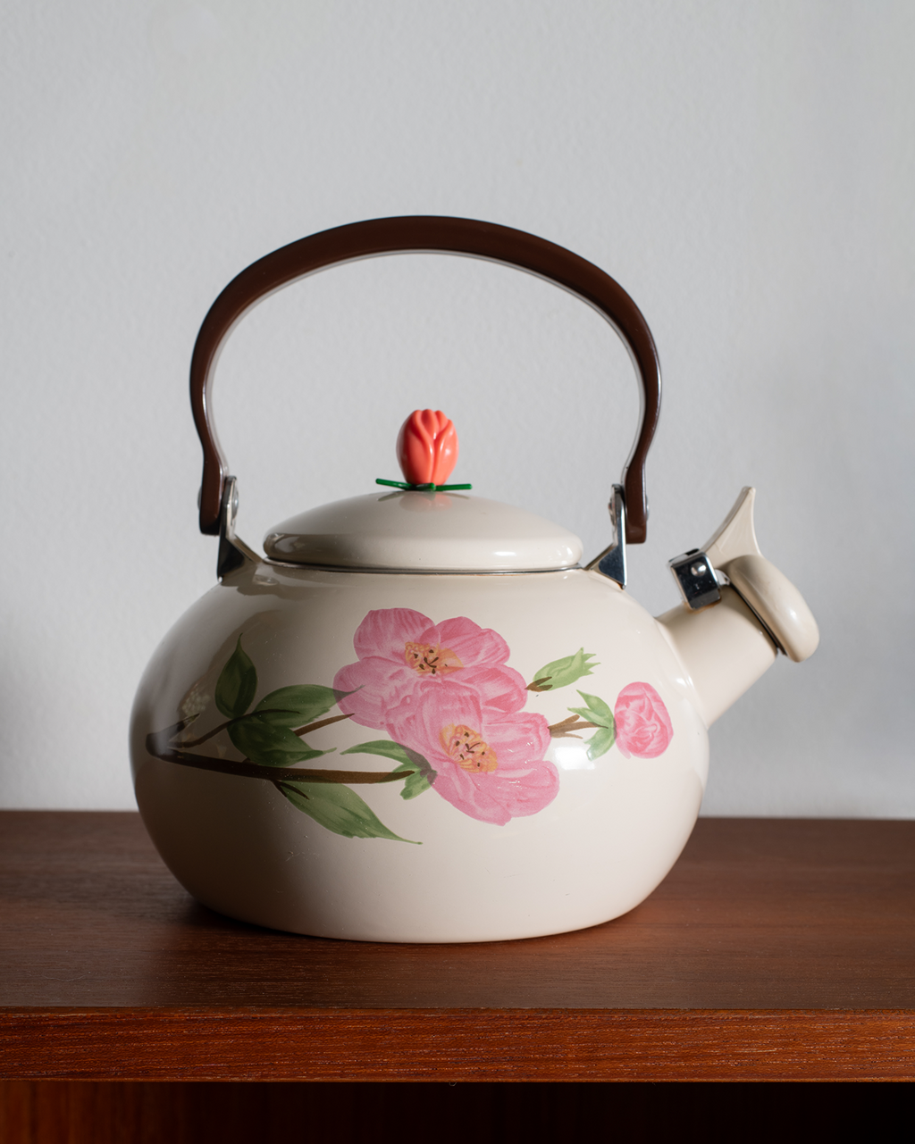 A cream-colored ceramic teapot with pink cherry blossom floral illustrations, a brown wooden handle, and a small pink tulip-shaped knob on the lid, resting on a wooden surface against a light gray background.