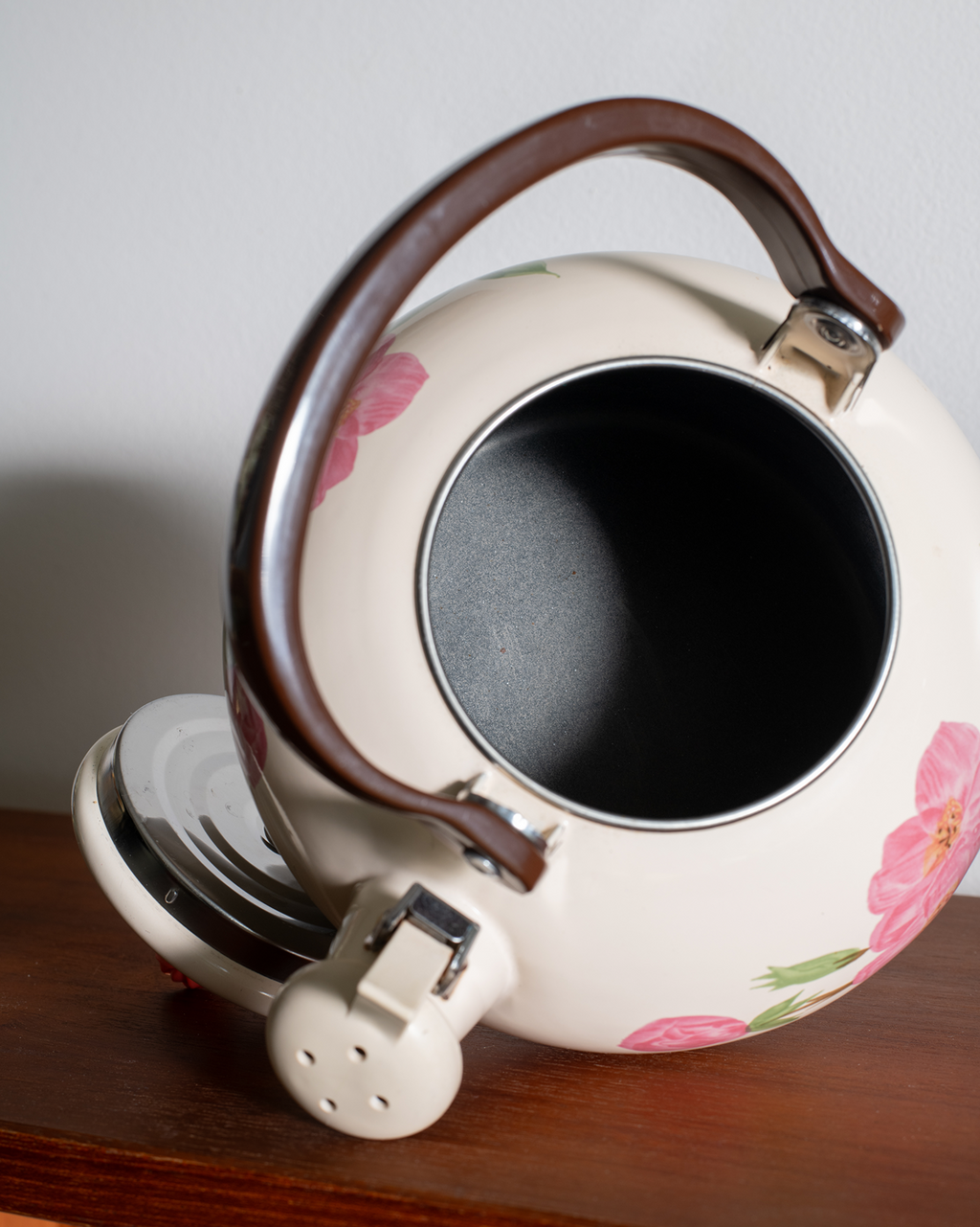 A cream-colored enamel tea kettle with pink floral patterns, a dark brown wooden handle, and silver metal accents, shown resting on a wooden surface against a neutral background.