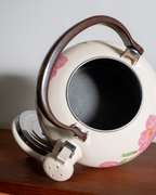 A cream-colored enamel tea kettle with pink floral patterns, a dark brown wooden handle, and silver metal accents, shown resting on a wooden surface against a neutral background.