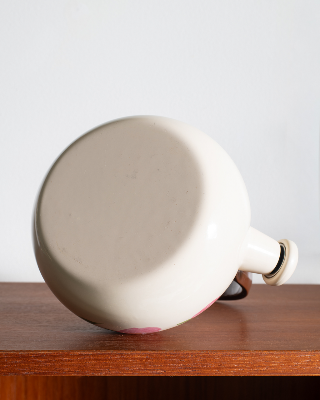 A vintage cream-colored enamel jug with a pink floral pattern visible along the bottom edge, resting on a wooden shelf against a plain light gray background.