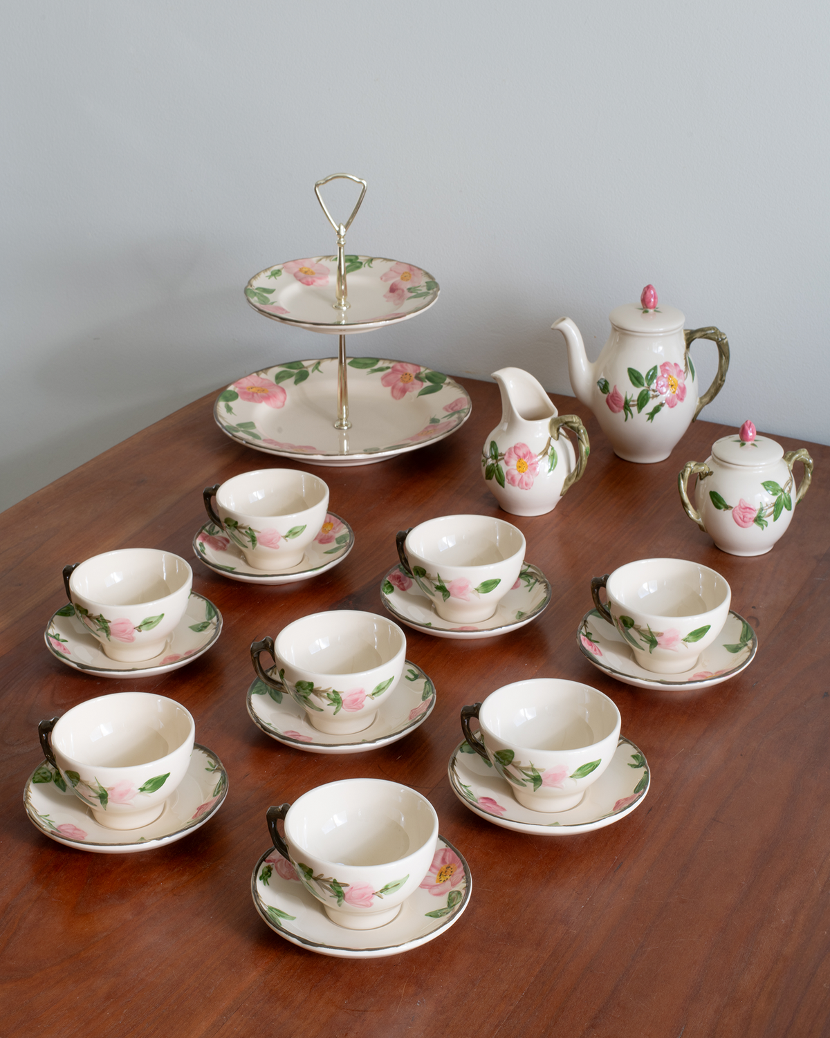 A vintage floral tea or coffee set arranged on a wooden table against a plain gray wall. The set includes multiple teacups and saucers with pink flowers and green leaves, a lidded sugar bowl, a creamer, a two-tiered serving stand, and a teapot.