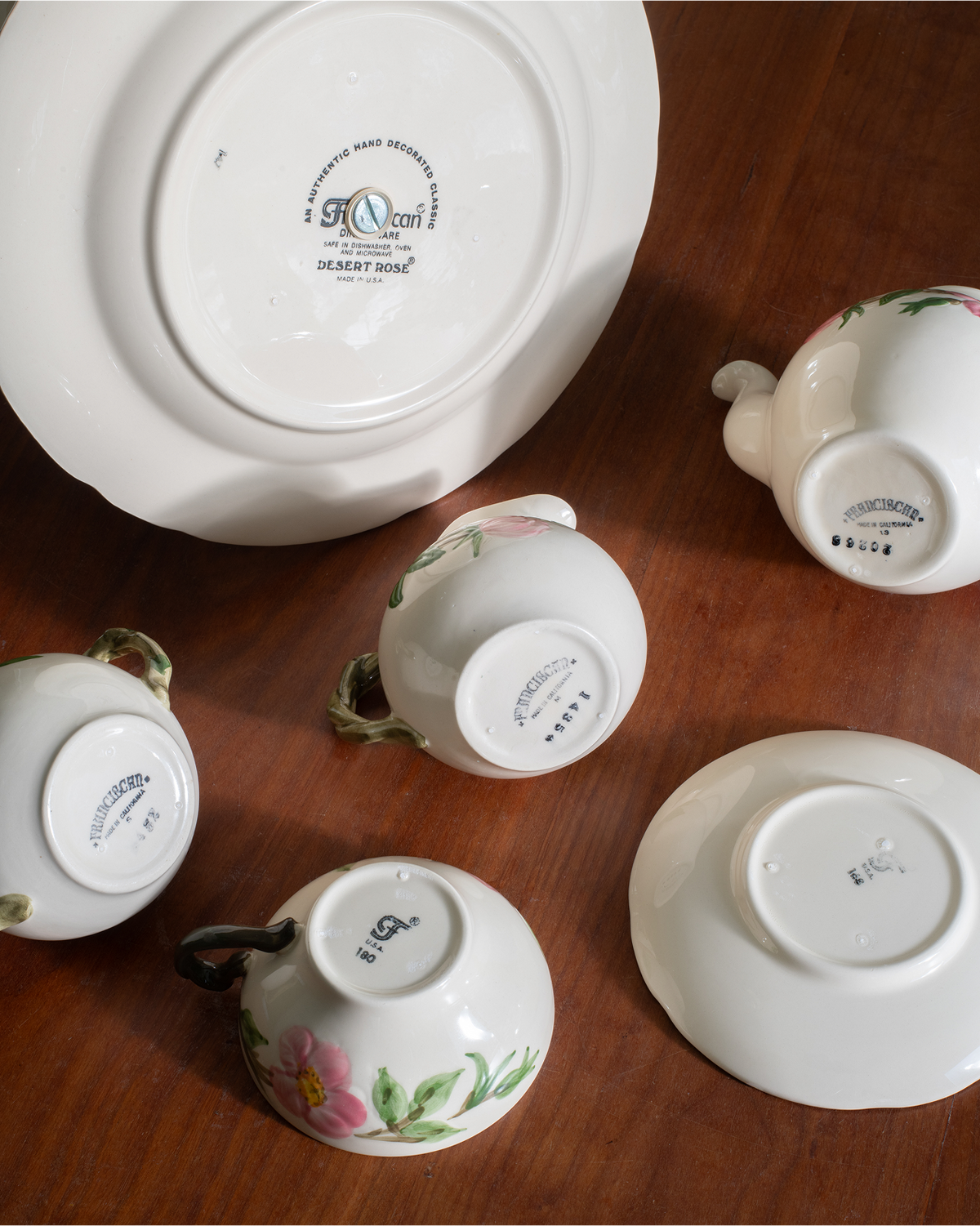 A set of white porcelain tableware pieces, including a large plate, teapot, cups, and saucers, arranged on a dark wooden surface. Several pieces are turned upside down to show the printed base markings, while one cup on the left is shown upright with its floral pink and green pattern visible on the exterior.