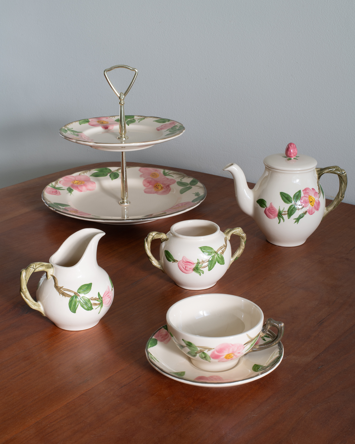 A ceramic tea and coffee set with a floral rose pattern, displayed on a wooden table against a gray background. The set includes a teapot, a creamer, a sugar bowl, a cup on a saucer, and a two-tiered serving stand with a gold-tone handle.