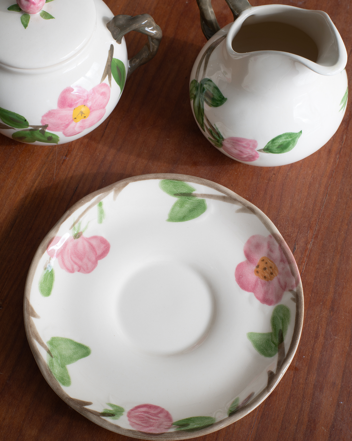 A white ceramic teapot and matching creamer pitcher with a floral saucer, decorated with pink flowers and green leaves, arranged on a dark wooden table.