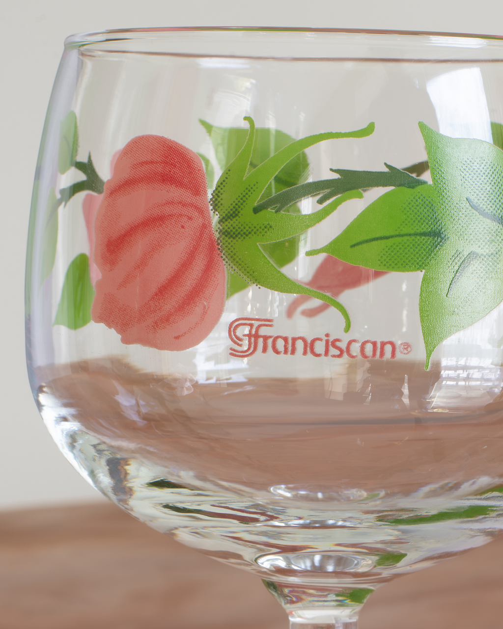 Close up of a clear wine glass with hand-painted pink flowers and green leaves, featuring the "Franciscan" logo, against a plain light wall.