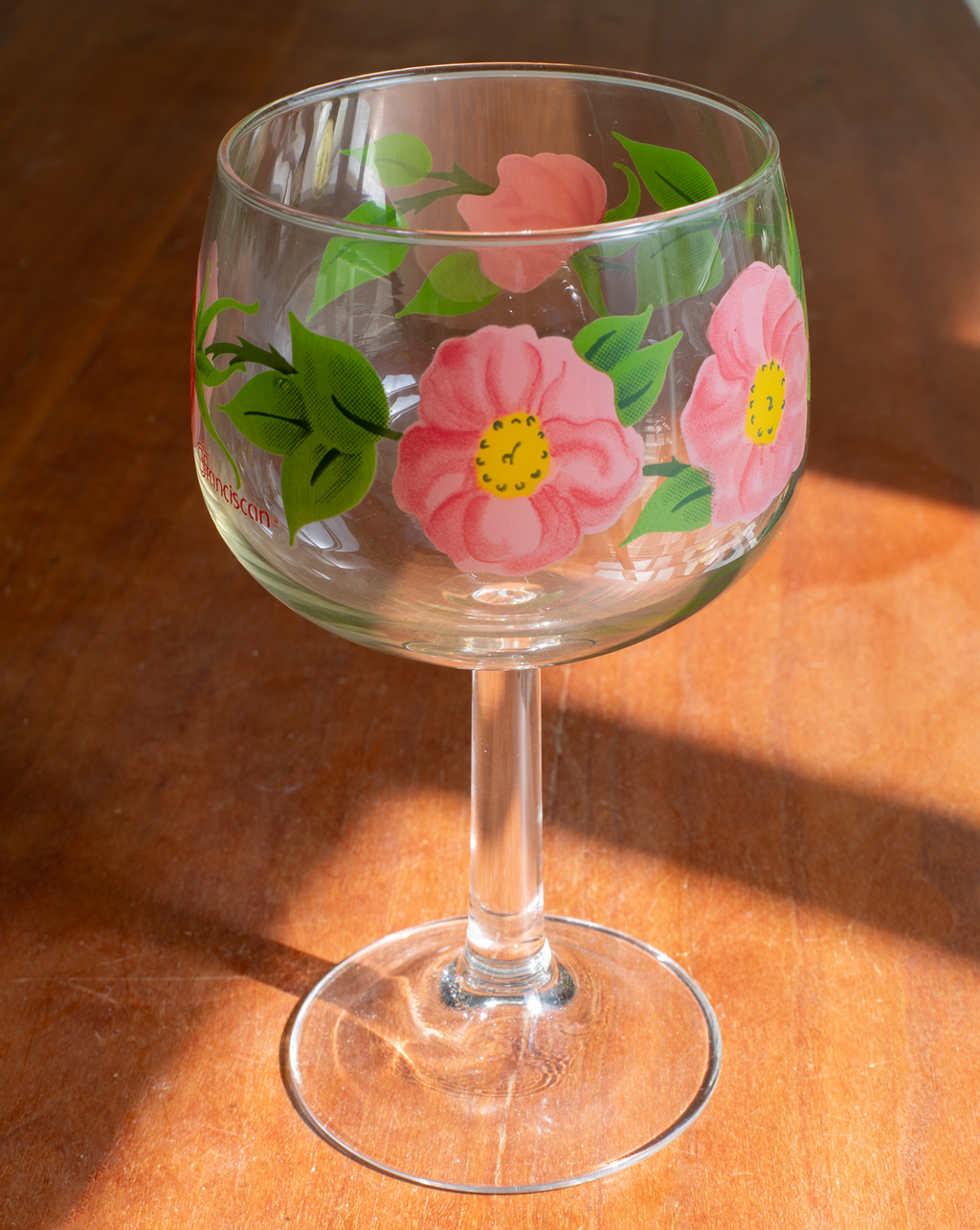 A clear stemmed wine glass decorated with hand-painted pink flowers and green leaves, resting on a wooden surface in warm sunlight.