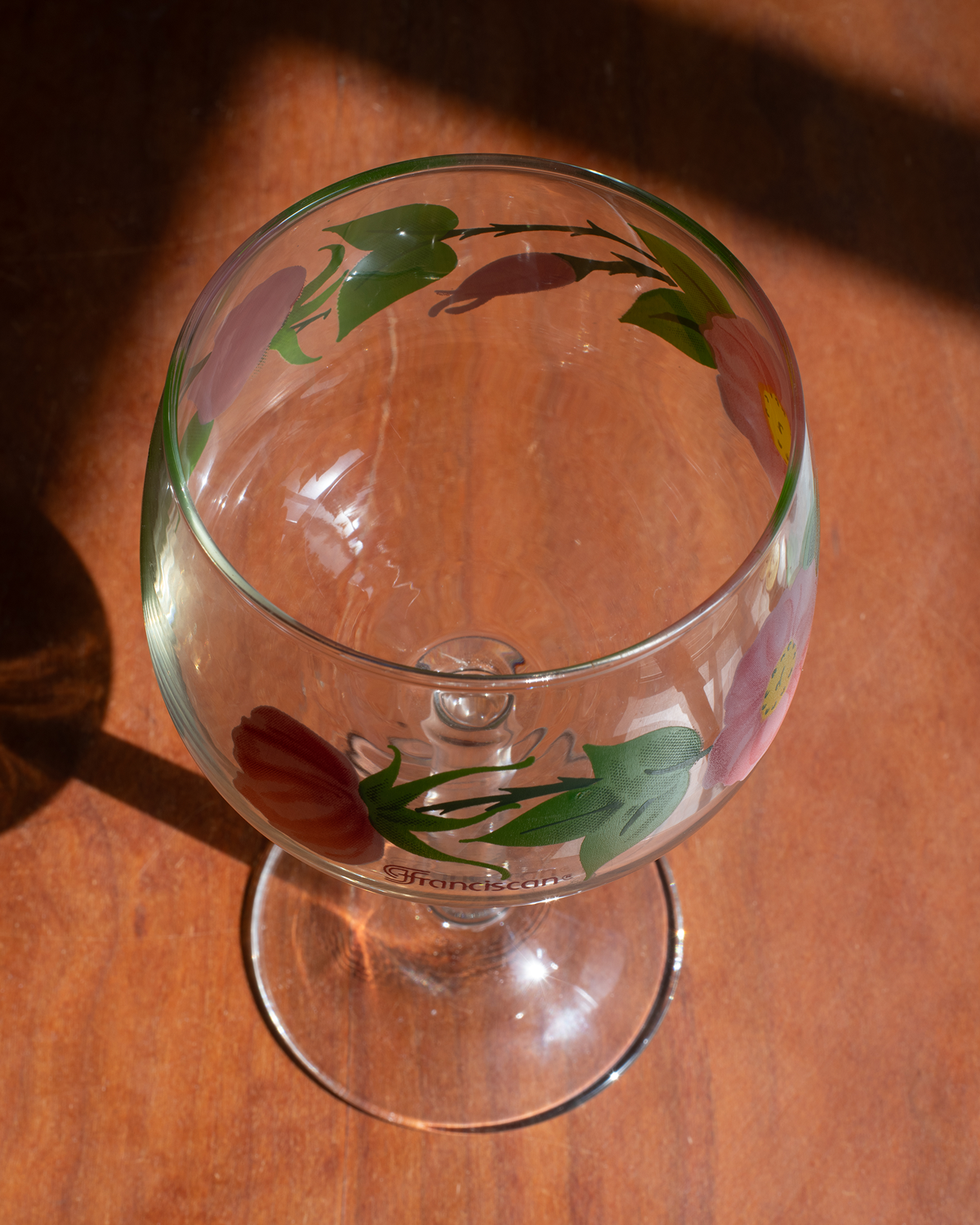 A clear glass goblet with hand-painted floral designs in green, pink, and red, resting on a warm wooden surface under direct sunlight.