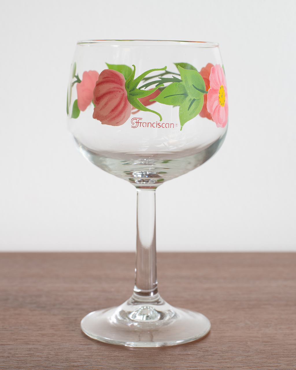 A clear glass wine glass with a floral design featuring pink flowers and green leaves, with the word 'Franciscan' printed on the side, set on a wooden surface against a white background.