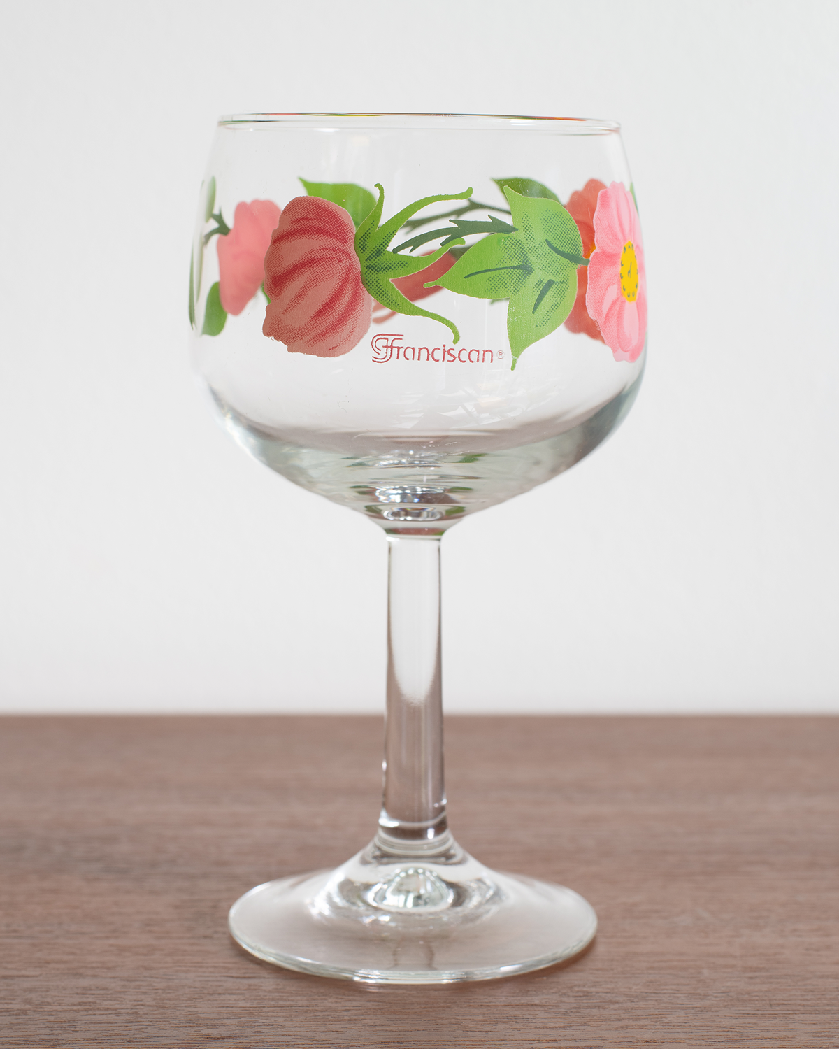 A clear glass wine glass with a floral design featuring pink flowers and green leaves, with the word 'Franciscan' printed on the side, set on a wooden surface against a white background.