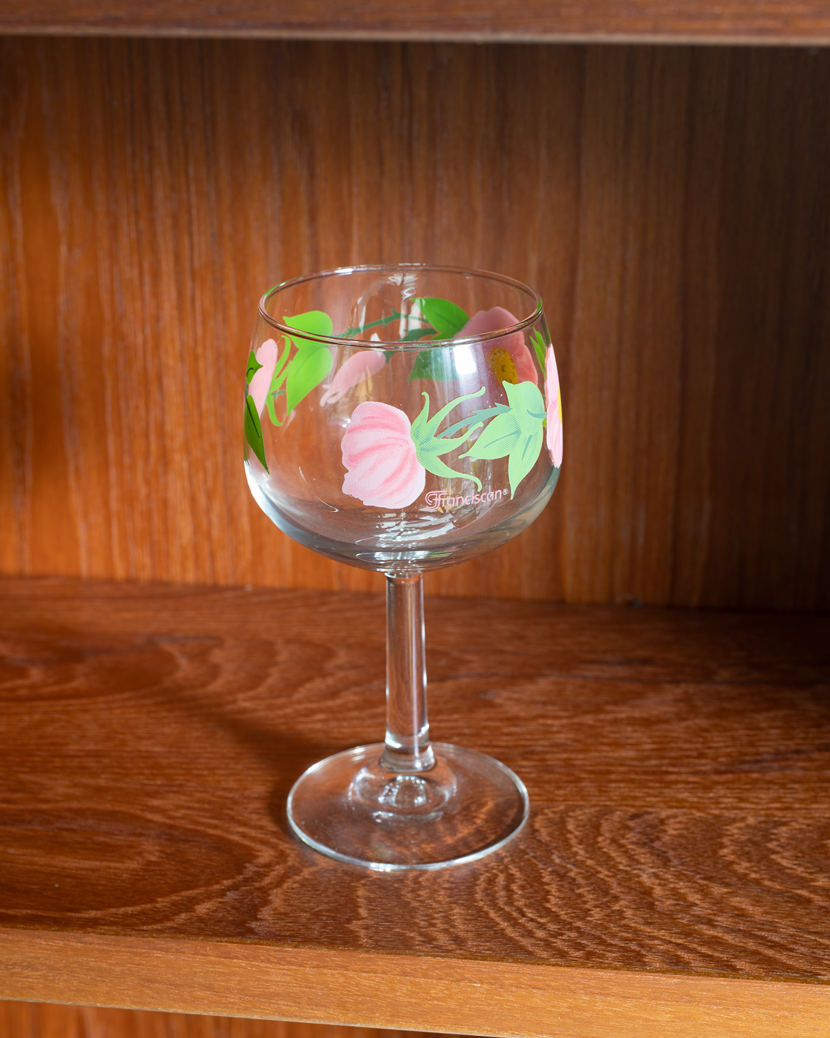 A clear glass wine glass with a stem, decorated with hand-painted pink flowers and green leaves, displayed on a wooden shelf.