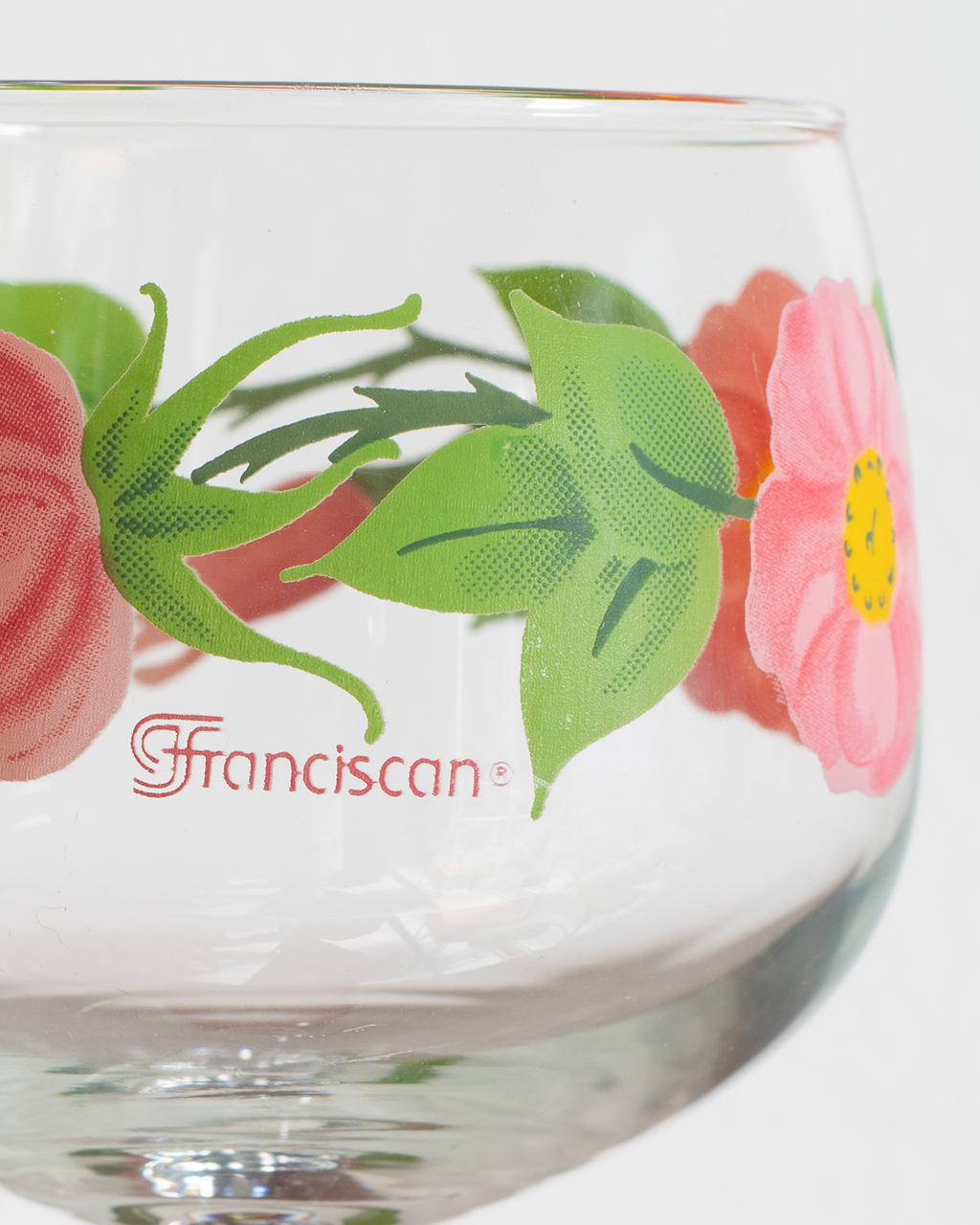 A close-up of a clear glass bowl or cup featuring a printed floral design with pink flowers, green leaves, and the word 'Franciscan' in red script near the bottom.