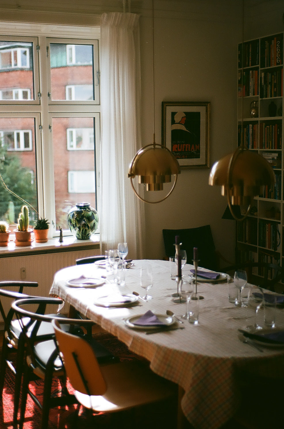 A dining room scene with a round table set for multiple guests, two hanging dome-shaped pendant lights, a window with daylight and outdoor view, chairs, a bookshelf, and framed wall art.
