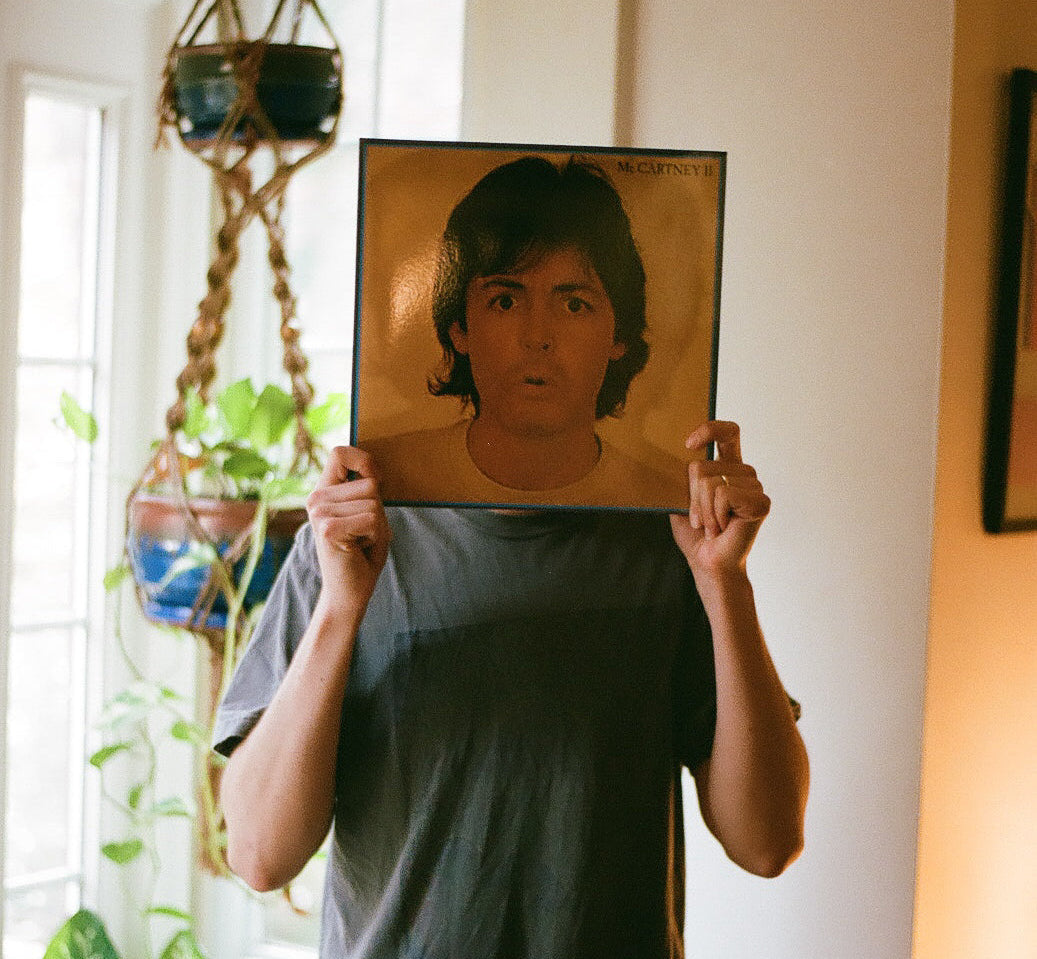 A person in a dark gray t-shirt holds up a framed portrait of Paul McCartney with a surprised expression, standing in a brightly lit room with houseplants and a macrame plant hanger visible.