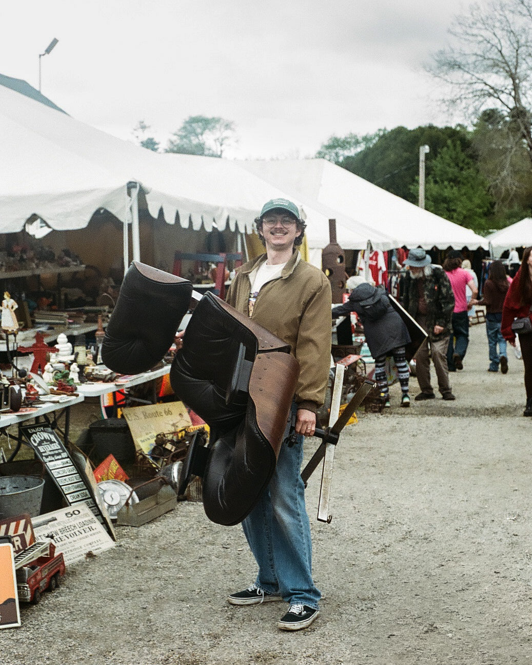 A person standing outdoors at Brimfield market or fair holding a large black Eames chair, with white tents, tables, and other people in the background.