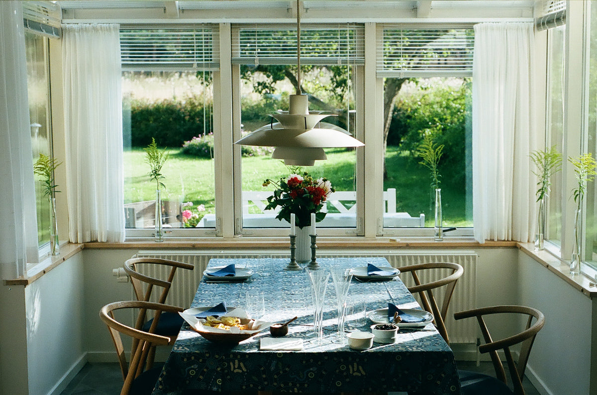 A Danish dining table set with a blue patterned tablecloth, blue napkins, and a vase of red flowers, surrounded by wooden chairs in a bright sunroom with large windows overlooking a green garden.
