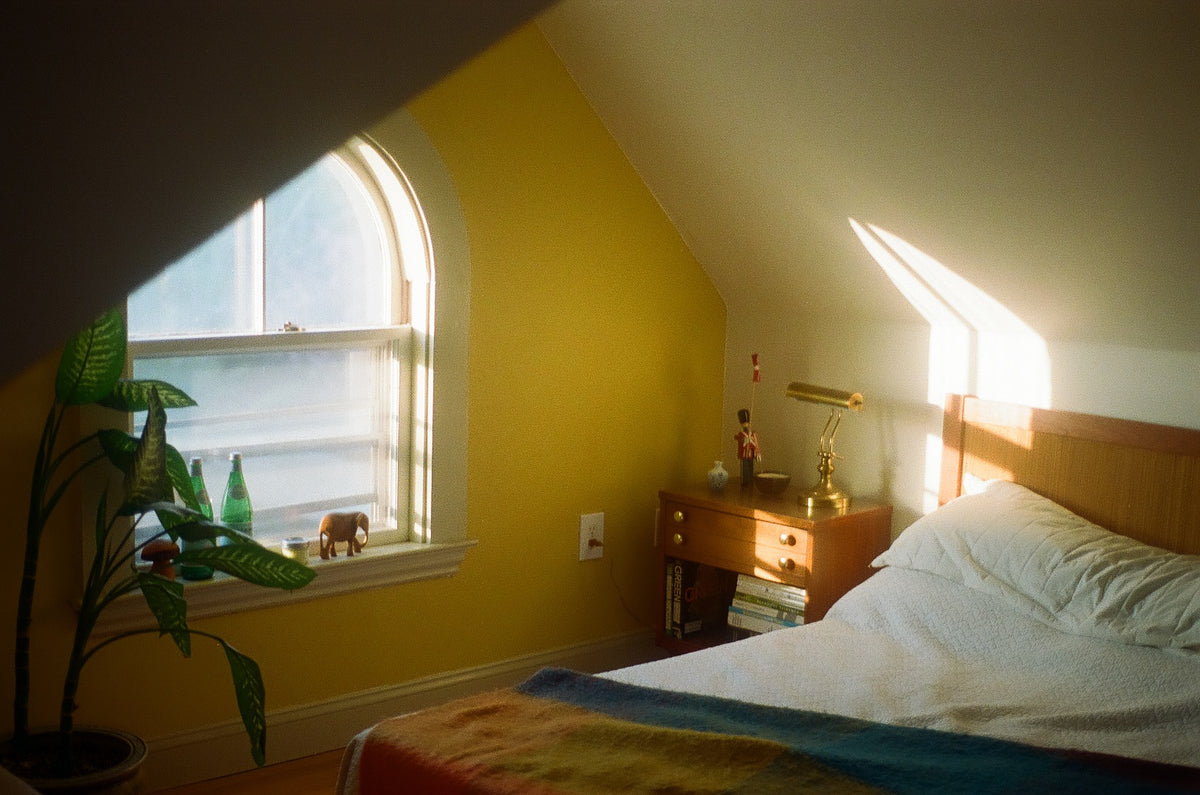 A cozy bedroom corner with a bed, wooden nightstand, and a bright yellow wall under a slanted ceiling. Sunlight streams in from an arched window, and a potted plant sits on the windowsill.