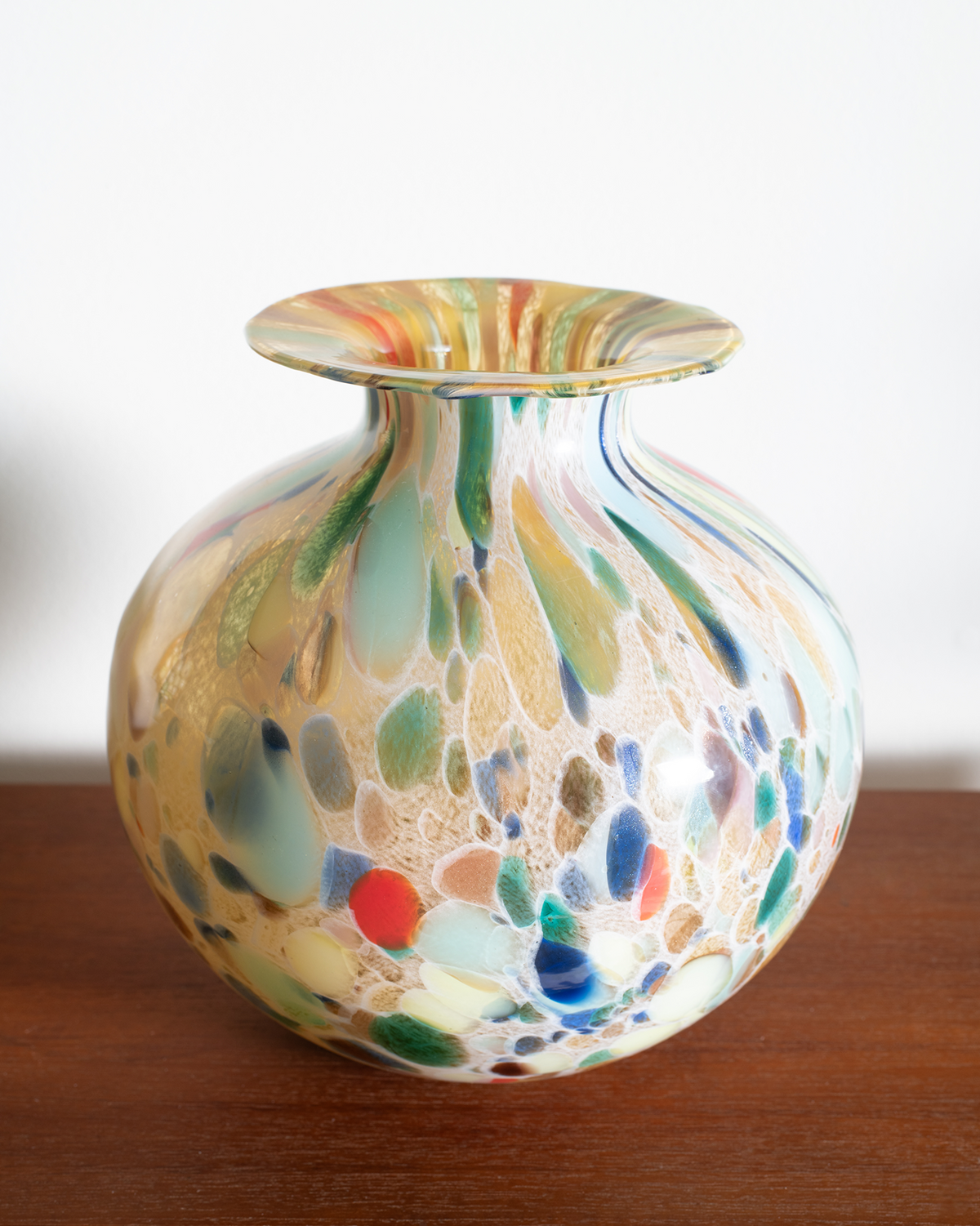 A hand-blown glass vase with a marbled pattern of beige, green, blue, red, and brown, sitting on a wooden surface against a plain white background.