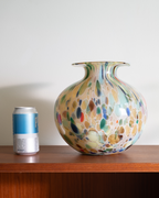 A colorful hand-blown glass vase with a rounded body and narrow neck, displayed on a wooden shelf next to a silver can for scale, against a plain light gray wall.