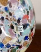 Close-up of a colorful glass decorative object with a mosaic pattern of blue, green, pink, brown, and white, featuring a small white 'MADE IN ITALY' sticker.