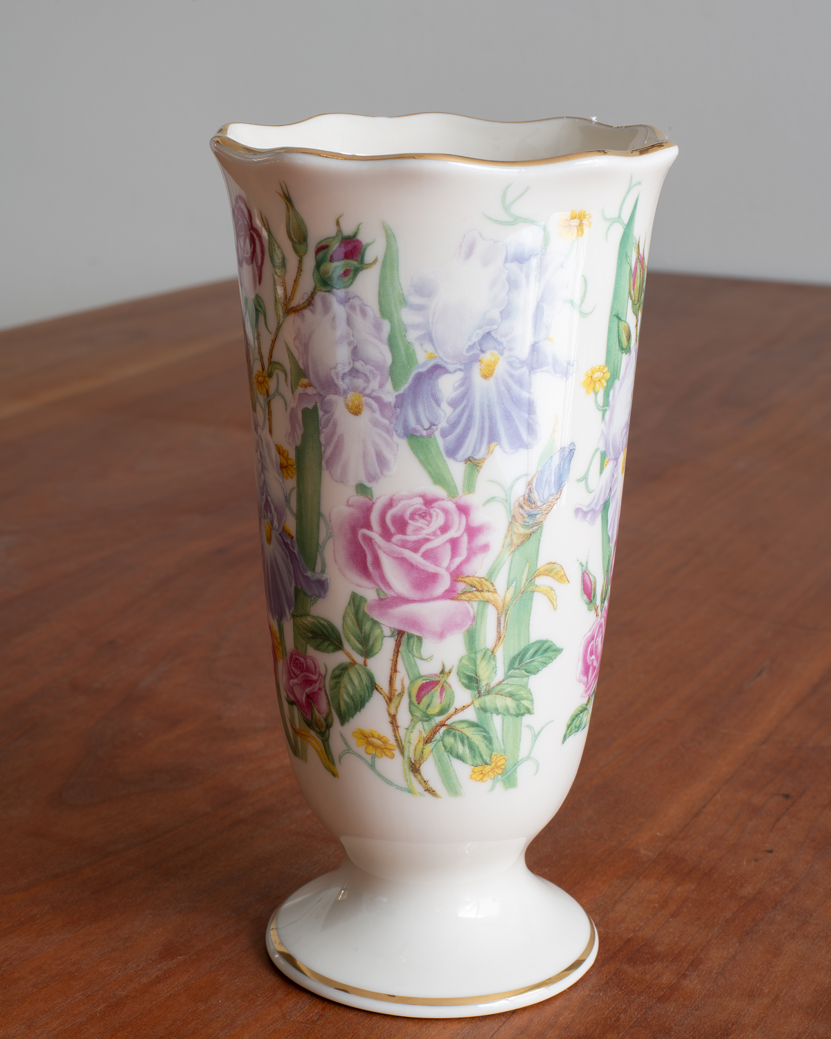 A white porcelain vase with a scalloped rim and pedestal base, decorated with a colorful floral pattern of pink roses, purple irises, and green leaves, with gold trim on the rim and base, set on a wooden table.