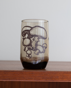 A translucent smoky-tinted glass tumbler with a dark brown mushroom illustration on the front, resting on a wooden surface against a light gray background.