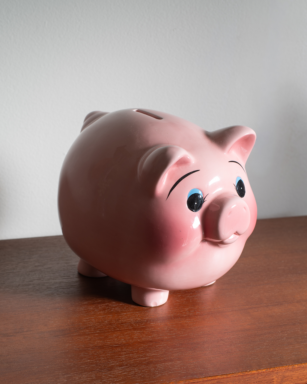 A glossy pink ceramic piggy bank with painted black eyebrows, blue eyes, and a pink snout, standing on a wooden table against a light gray wall.