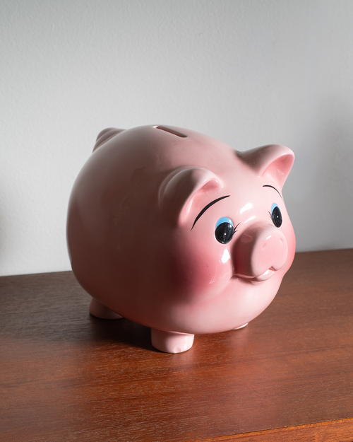 A glossy pink ceramic piggy bank with painted black eyebrows, blue eyes, and a pink snout, standing on a wooden table against a light gray wall.