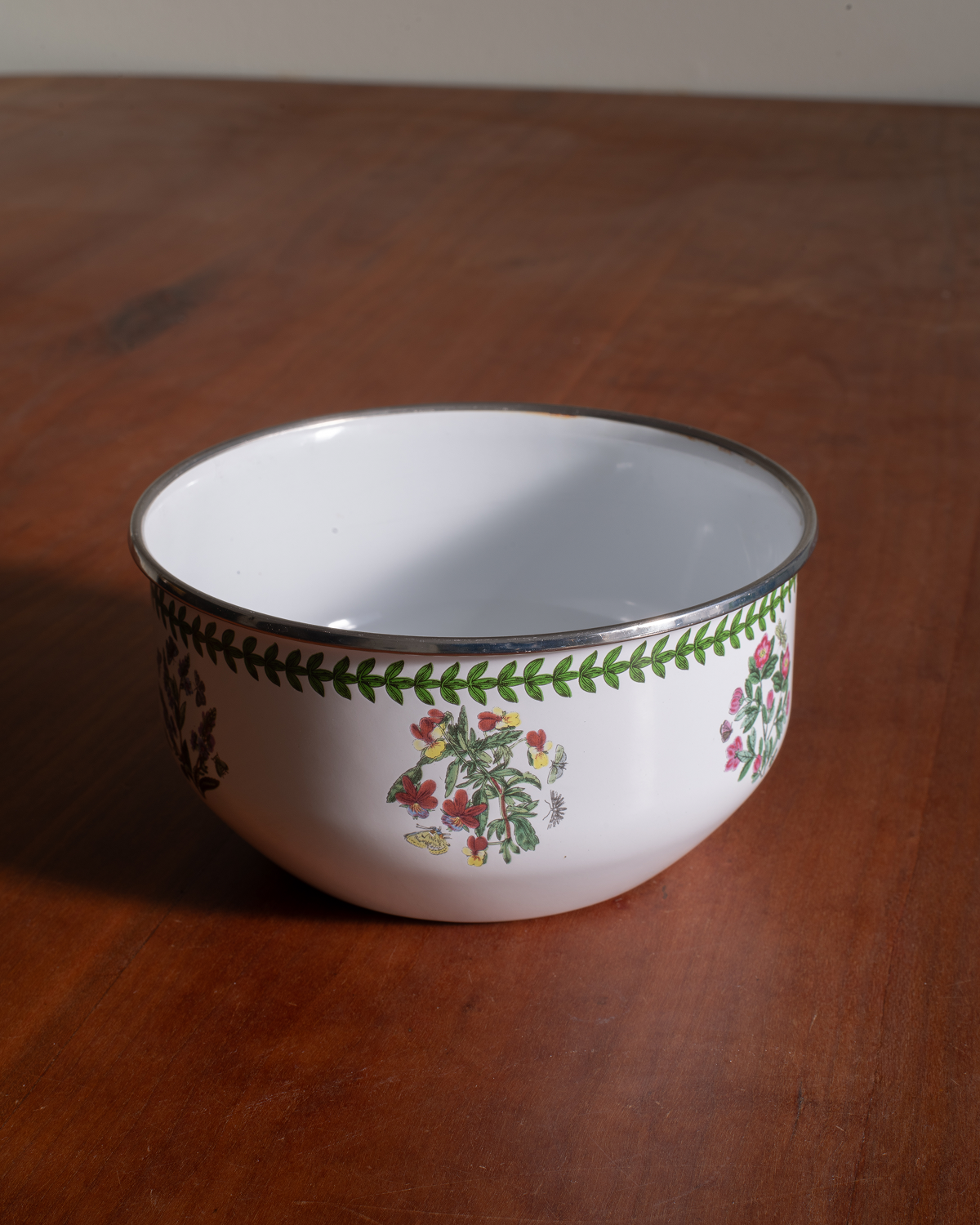 A white enamel bowl with a silver rim, decorated with green leaf patterns and small red, yellow, and blue floral motifs, shown on a wooden table.