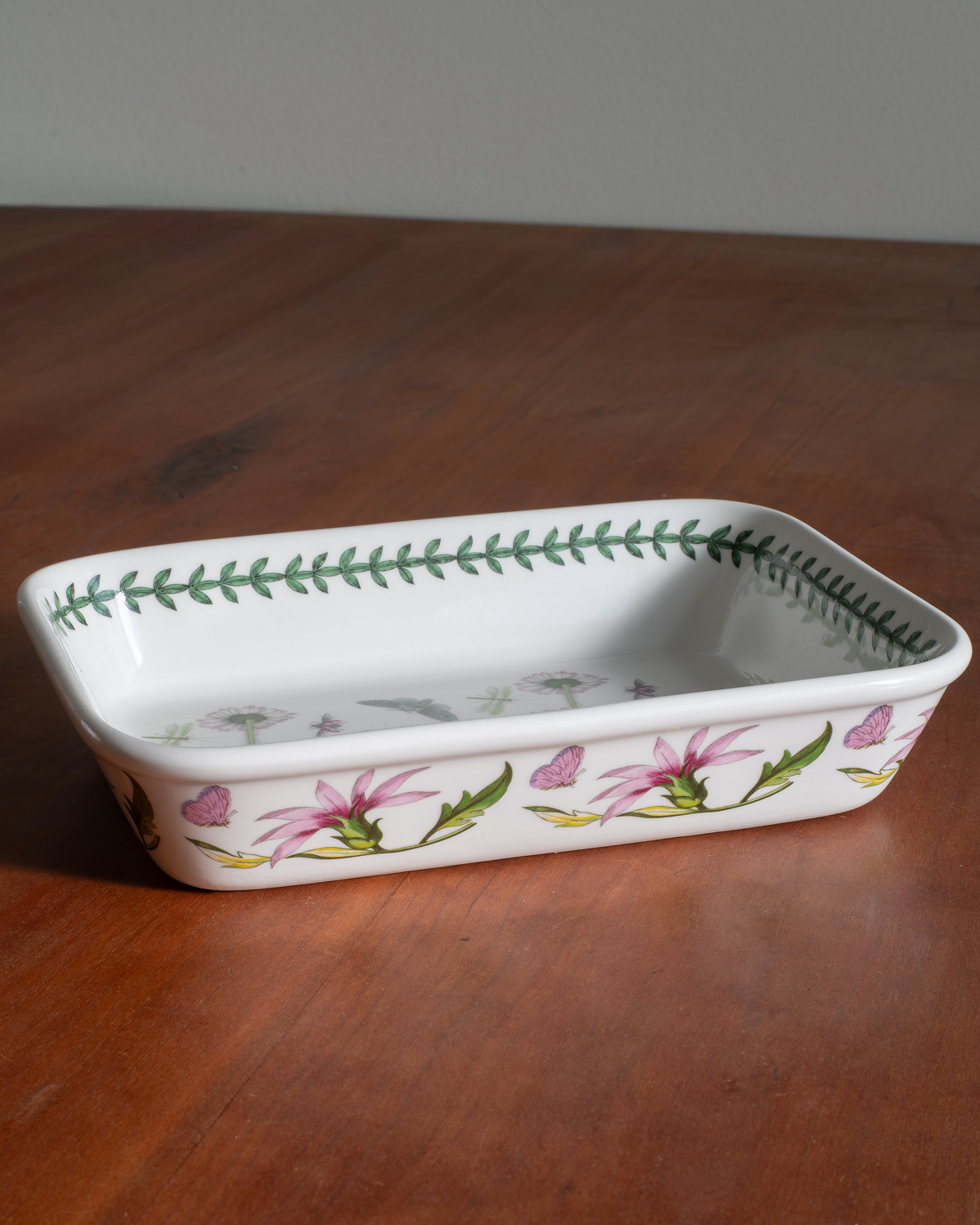 A rectangular white ceramic baking dish with a green leaf border on the inside and pink flowers with green leaves on the outside, resting on a wooden table.