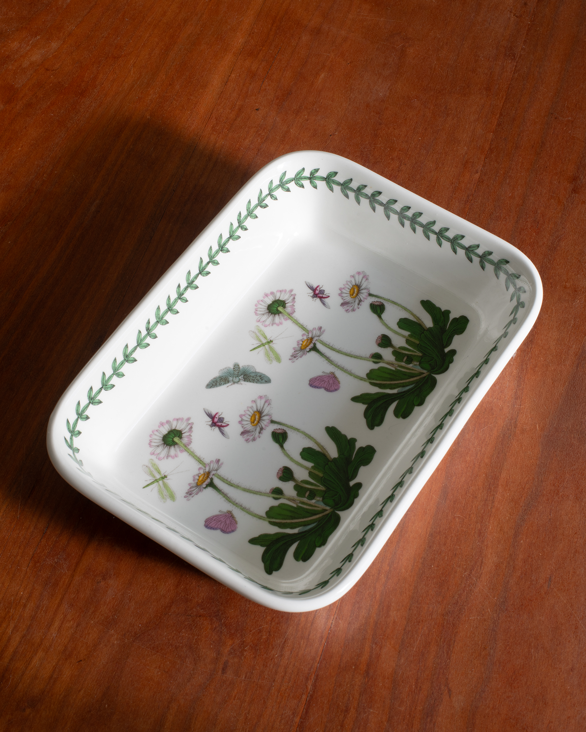 A white rectangular ceramic dish with rounded corners, featuring a green leaf border and delicate botanical illustrations of flowers and leaves inside, shown on a wooden table.