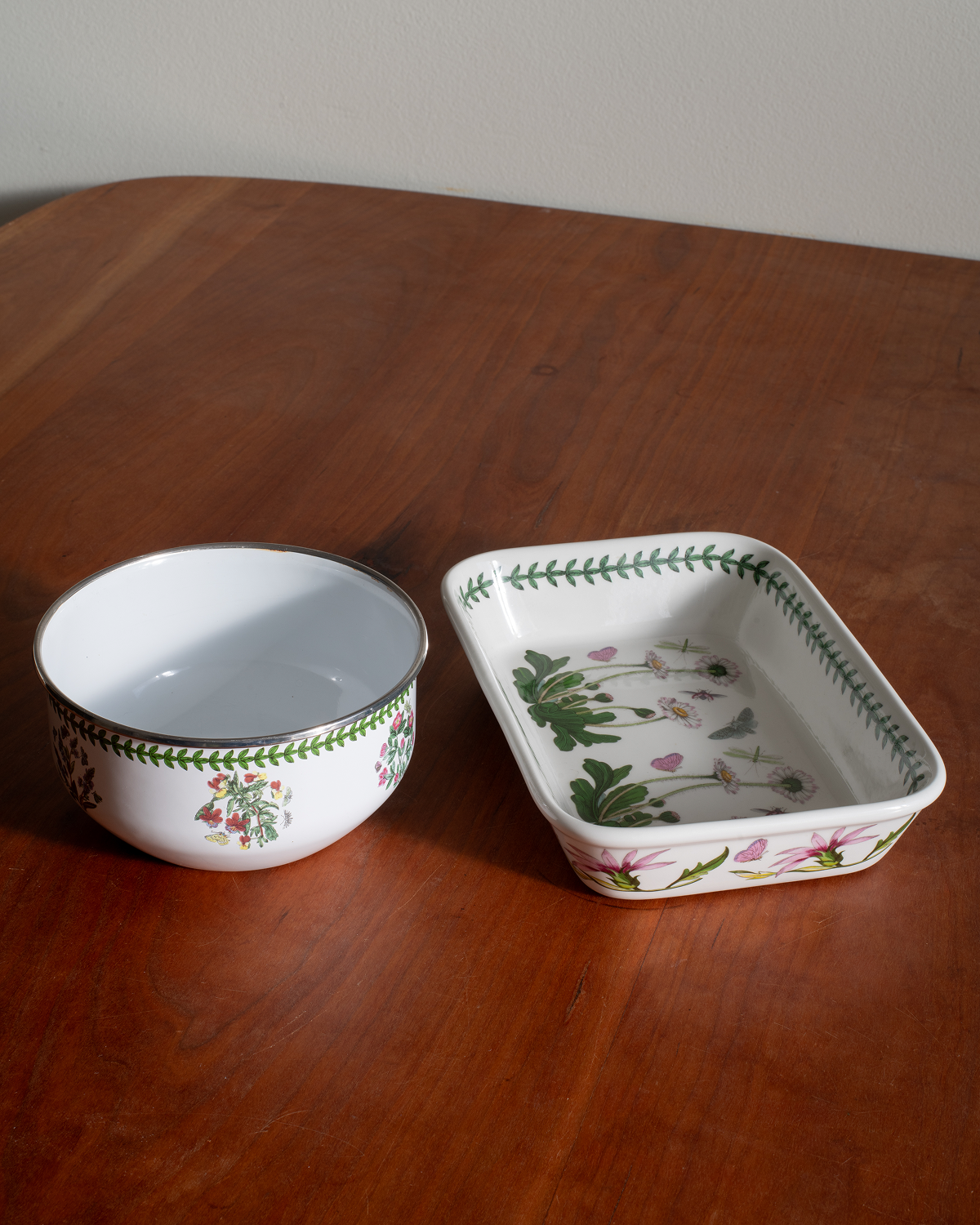 A white enamel bowl and matching rectangular baking dish with green botanical border patterns and pink floral details, displayed on a wooden table.