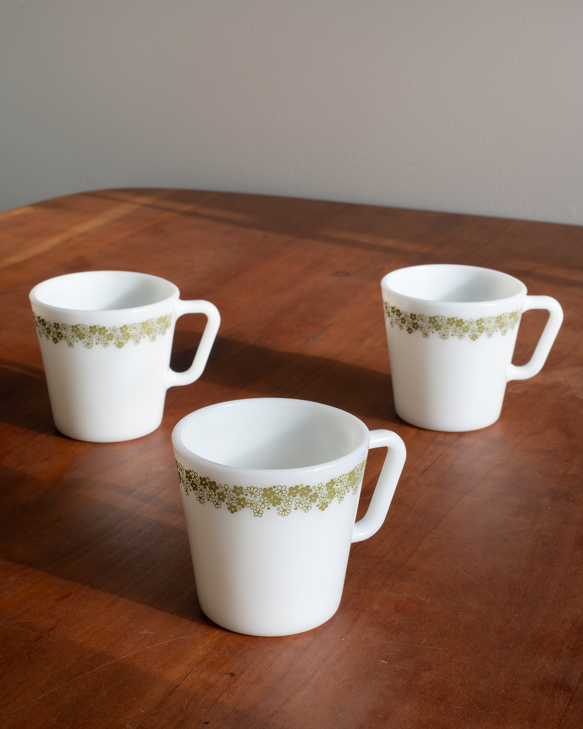 Three white mugs with a delicate olive green floral pattern band, arranged on a polished wooden table against a neutral gray background.
