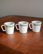 Three white coffee mugs with a green floral border pattern, arranged on a wooden table against a plain gray wall.