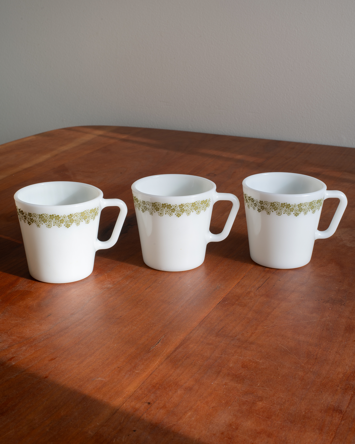 Three white mugs with a green floral pattern band, arranged side-by-side on a wooden table against a plain wall.
