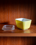 A square yellow ceramic bowl with a white interior sits on a wooden shelf next to a small clear plastic food tray against a dark wood-paneled background.