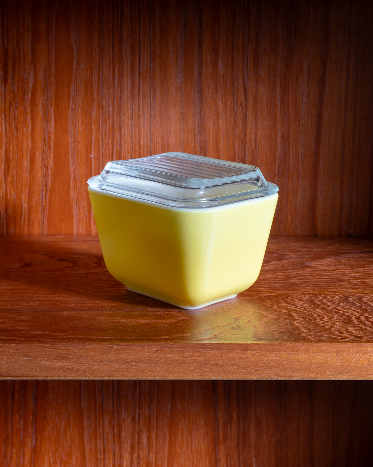 A small square yellow container with a clear ribbed plastic lid, sitting on a wooden shelf against a dark wood background.