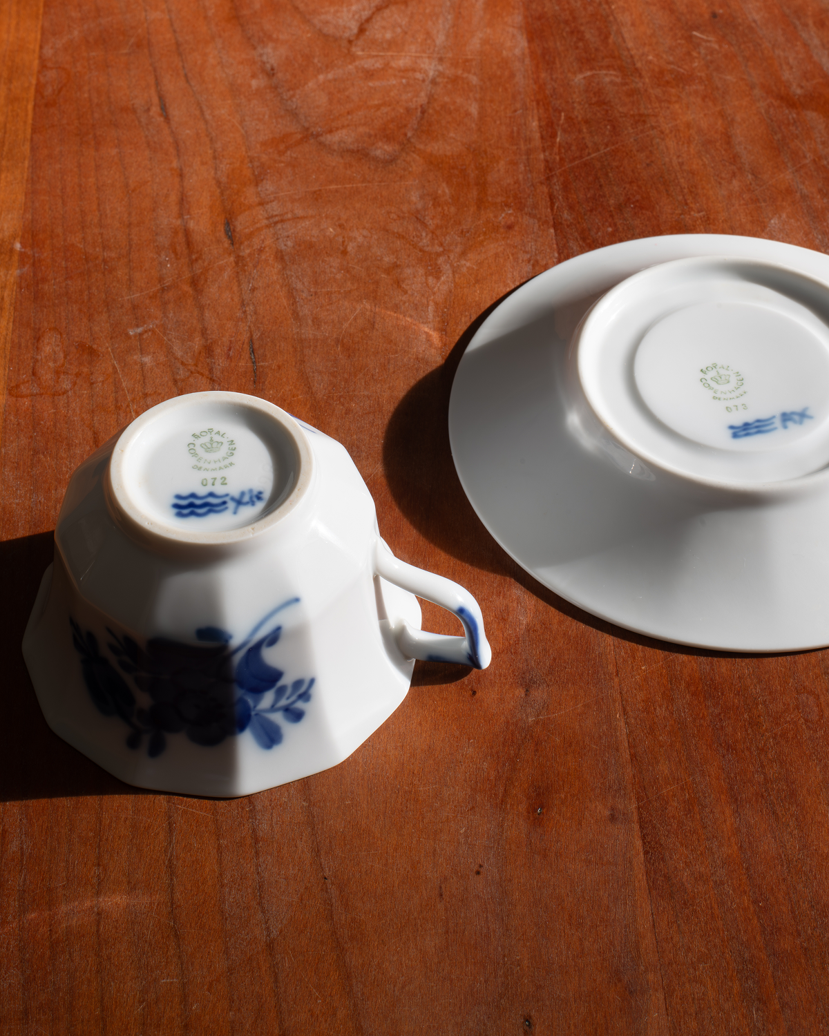 A white porcelain teacup and saucer set with blue floral decoration, photographed upside down on a warm brown wooden table in direct sunlight.