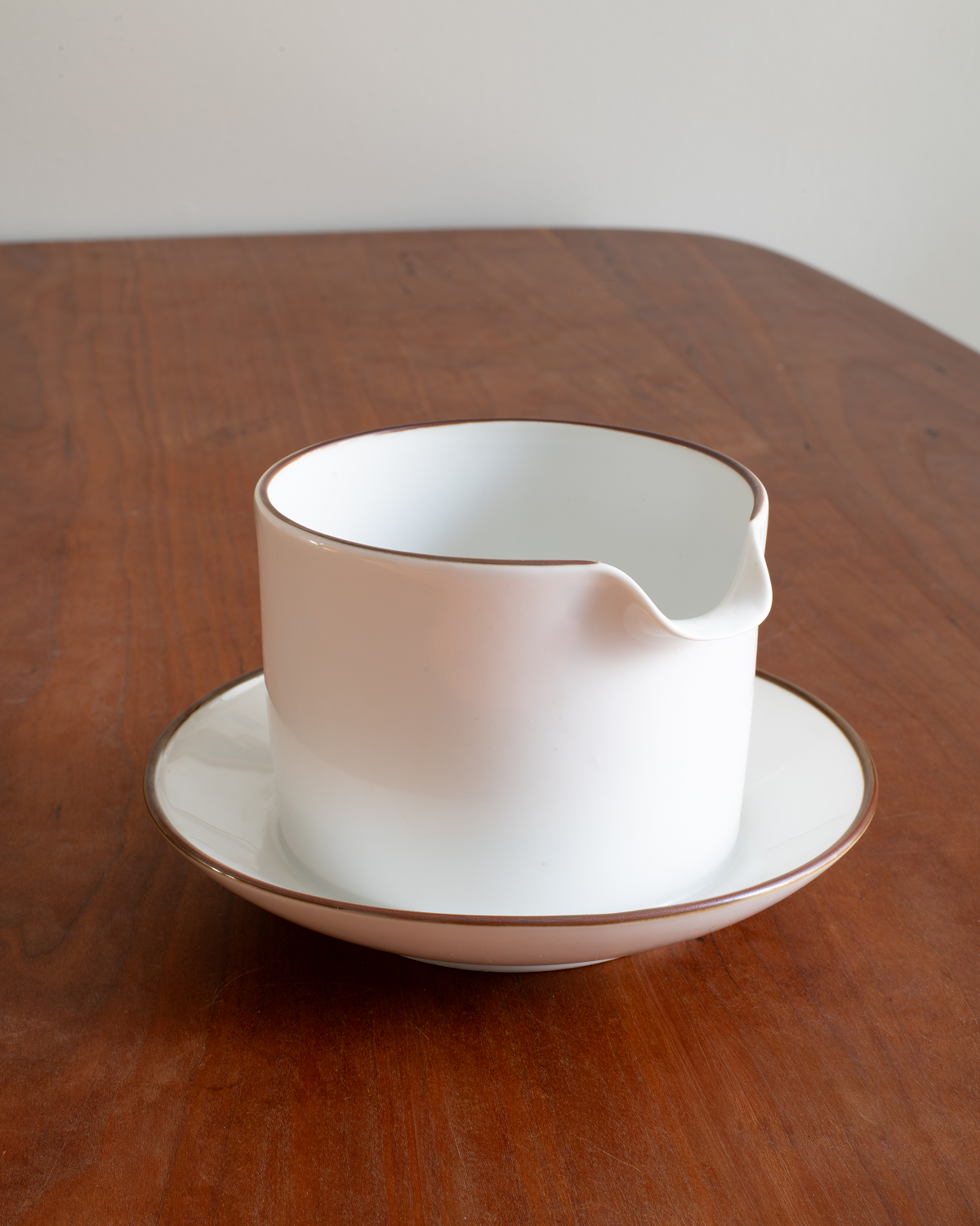 A white ceramic pitcher with a small pouring spout and matching saucer, both accented with a thin brown rim, set on a dark wood table.
