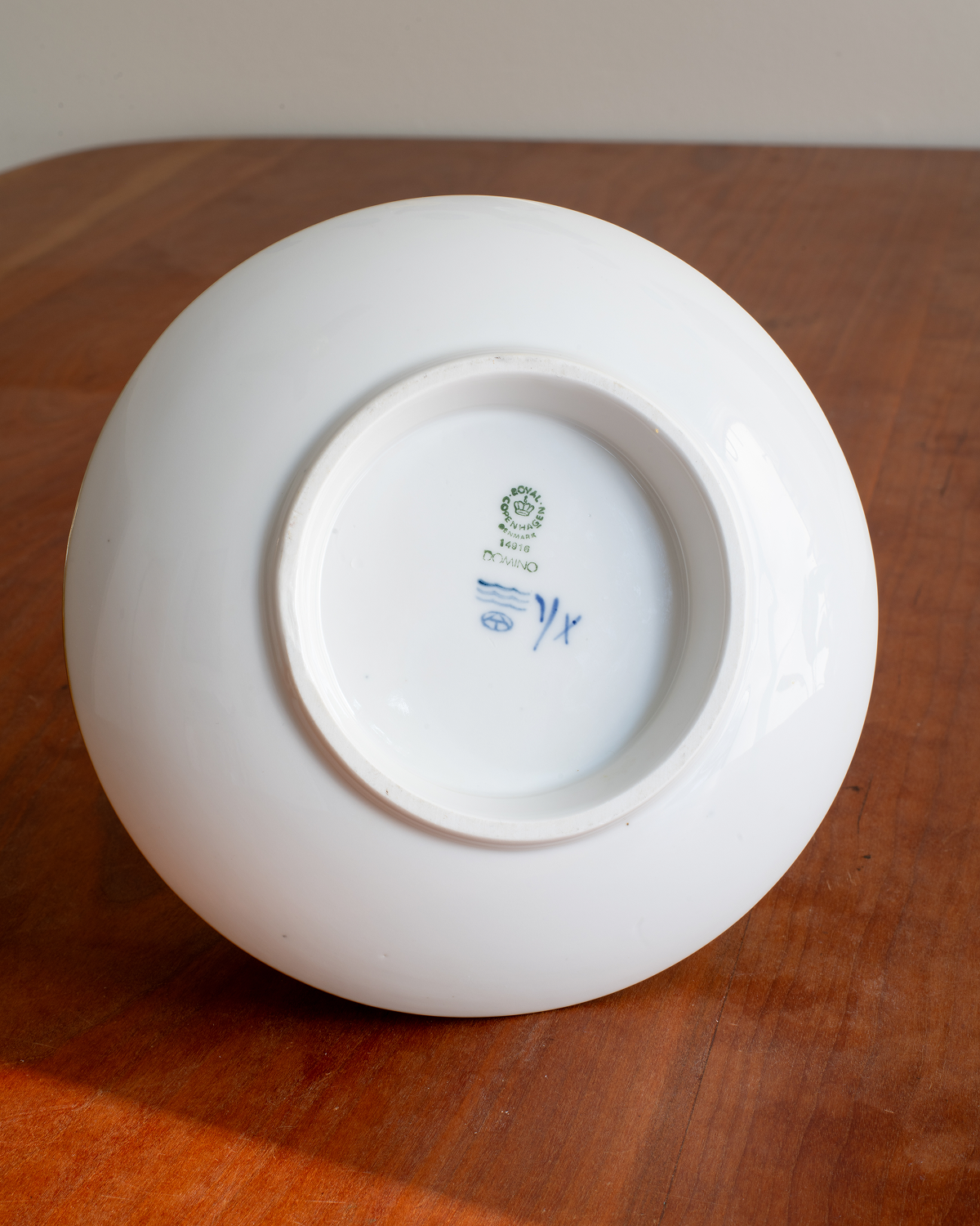 The bottom of a glossy white ceramic bowl or plate resting on a warm brown wooden surface, showing the printed blue and green manufacturer's mark inside the base.