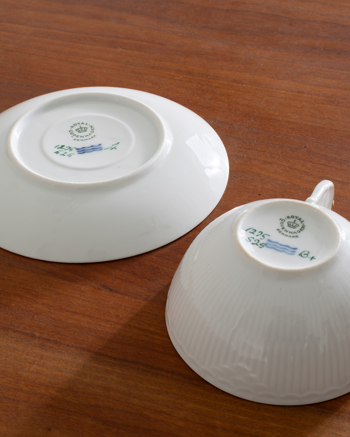 Two white ceramic teacups shown upside down on a wooden table, displaying their printed inner bottoms with a circular blue and green logo.