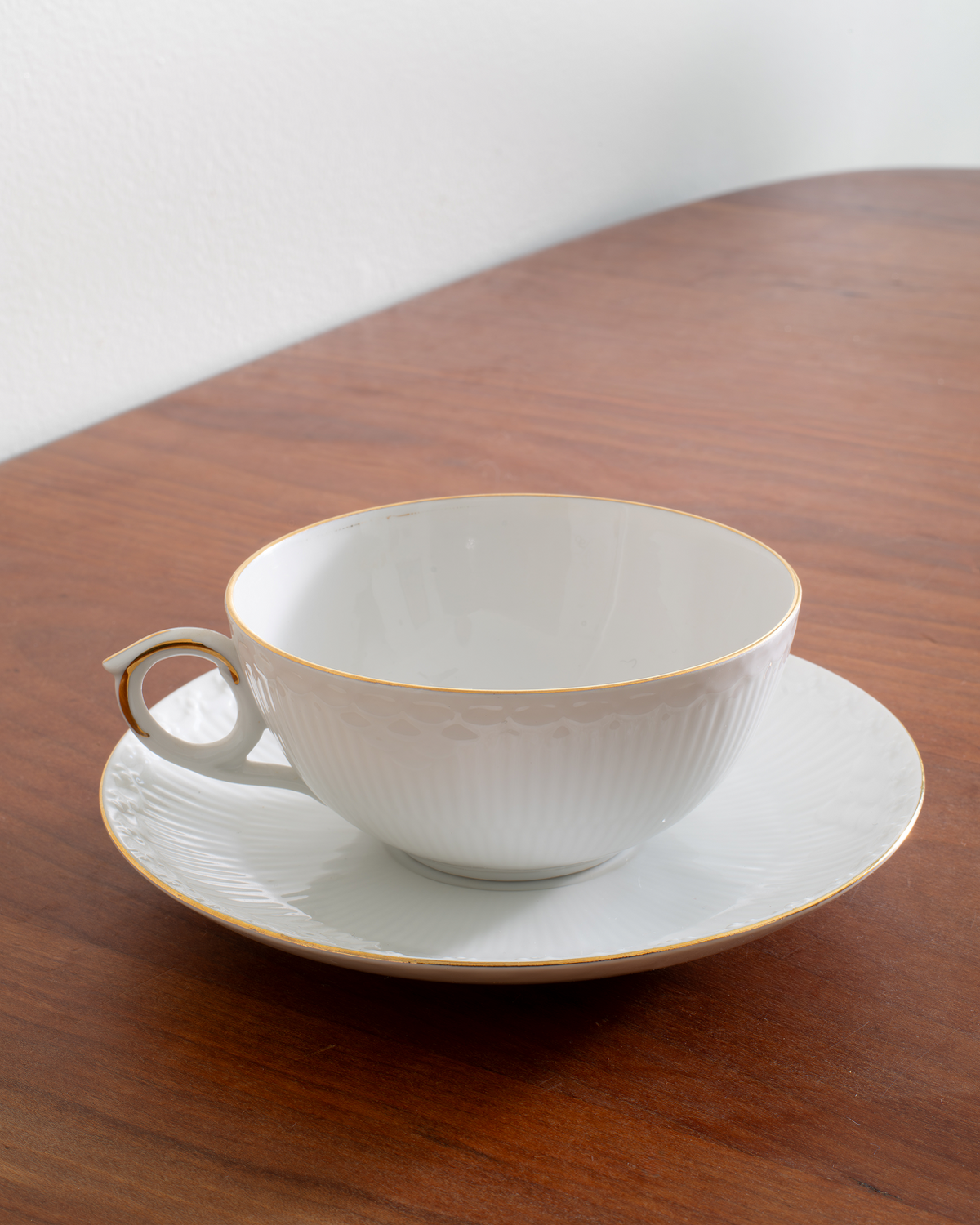 A white porcelain teacup and saucer set with delicate gold trim, placed on a warm brown wooden table against a light gray wall.