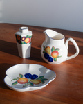 A white ceramic fruit-themed tableware set on a wooden table, including a small vase, a pitcher, and a shallow scalloped serving dish, all decorated with painted fruit motifs.