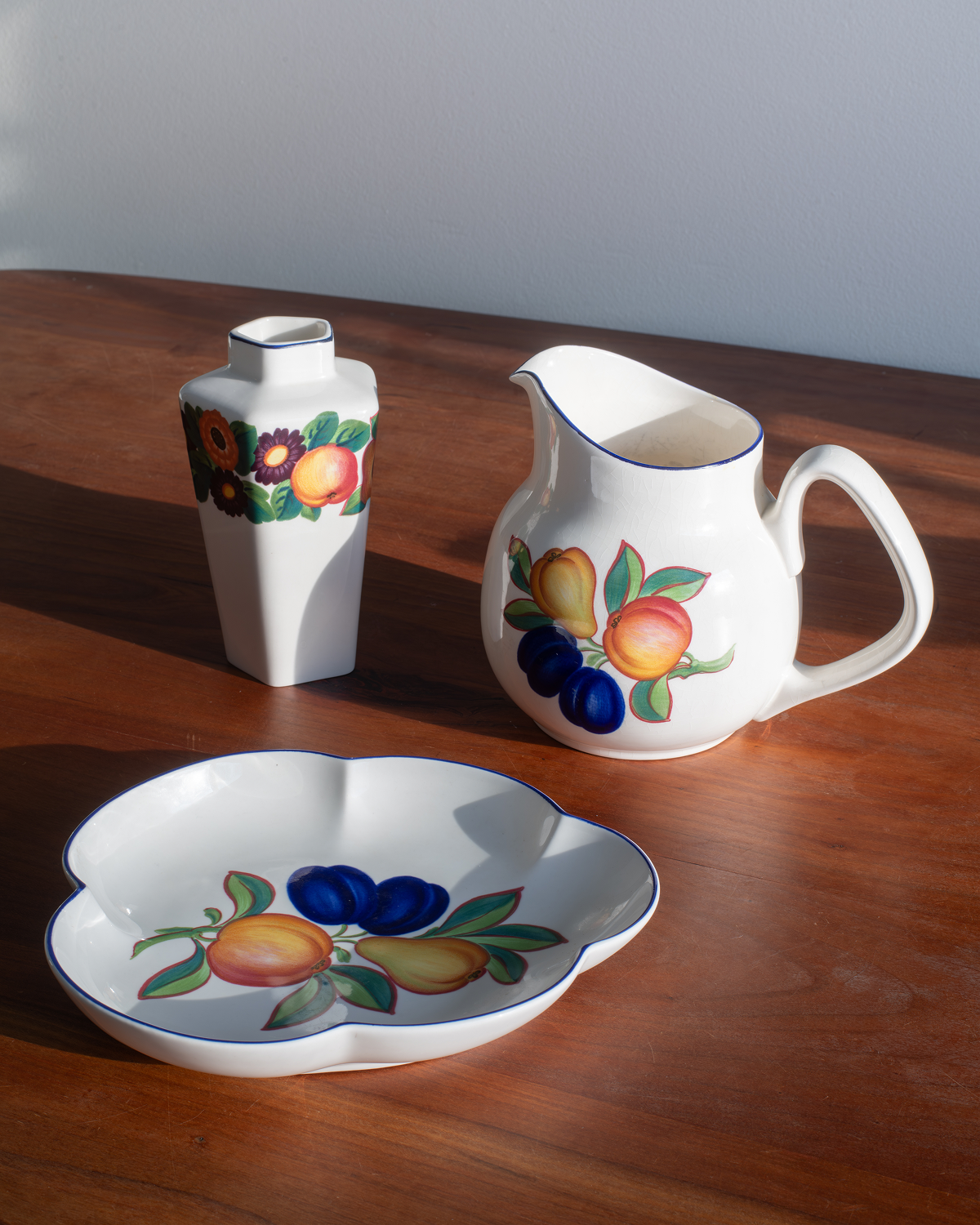 A white ceramic fruit-themed tableware set on a wooden table, including a small vase, a pitcher, and a shallow scalloped serving dish, all decorated with painted fruit motifs.