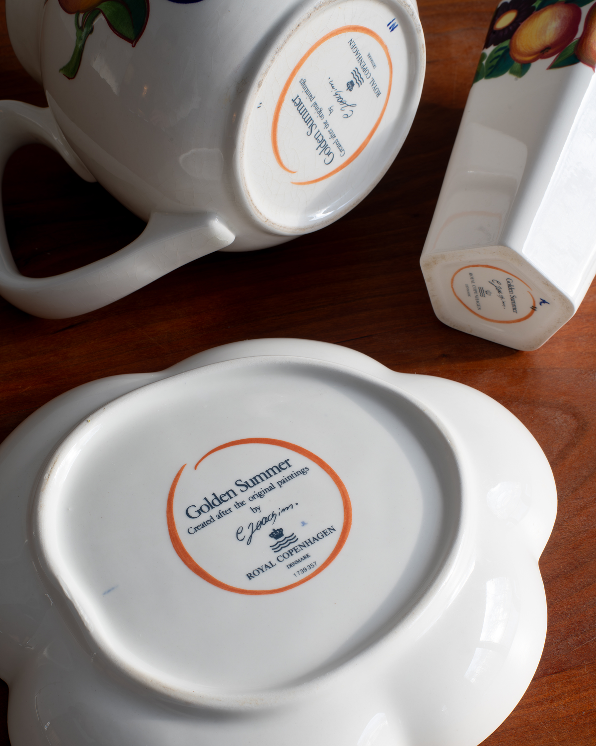White ceramic tableware pieces on a wooden surface, including a mug, a rectangular dish, and a plate, each showing the underside with a printed circular label. The center label reads 'Golden Summer' and 'Royal Copenhagen'.